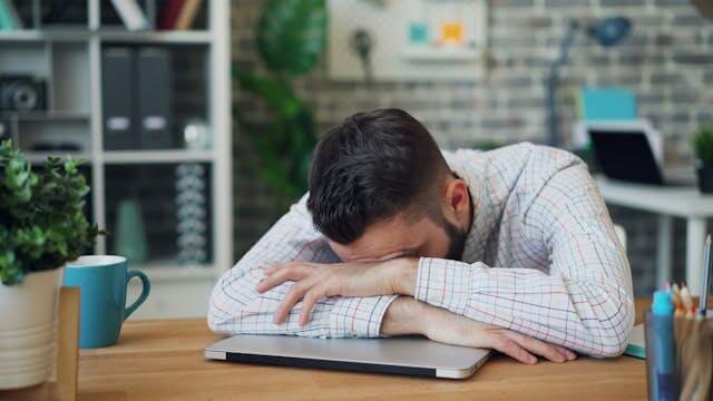 A man resting his head on a laptop, embodying signs you need better sleep in a home office setting.
