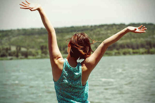 A woman embracing nature with open arms practicing holistic wellbeing rituals by the water.