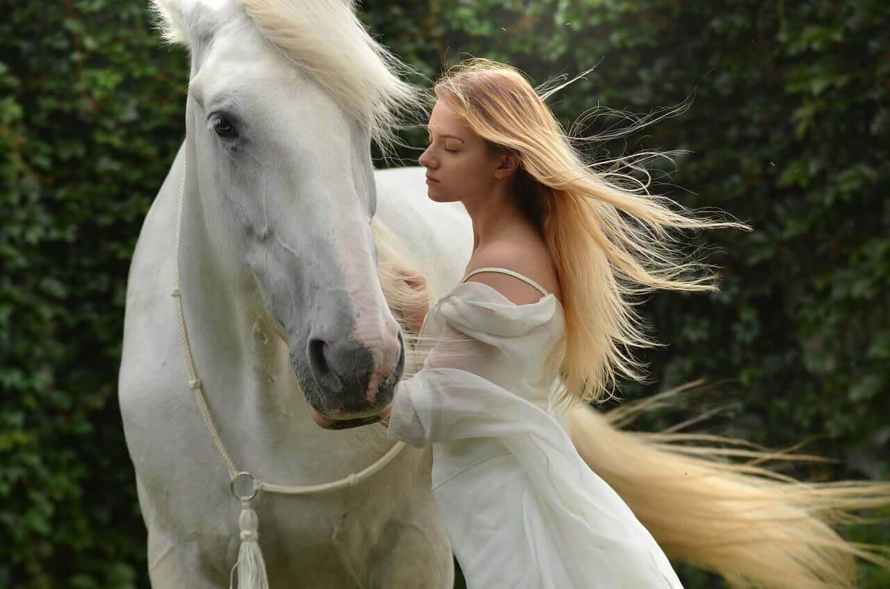 Woman in flowing white dress softly interacting with a white horse in a lush green setting embodying self-care products for women